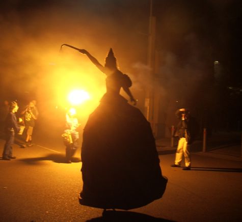 Woman on stilts illuminated by a flare, with whip raised.