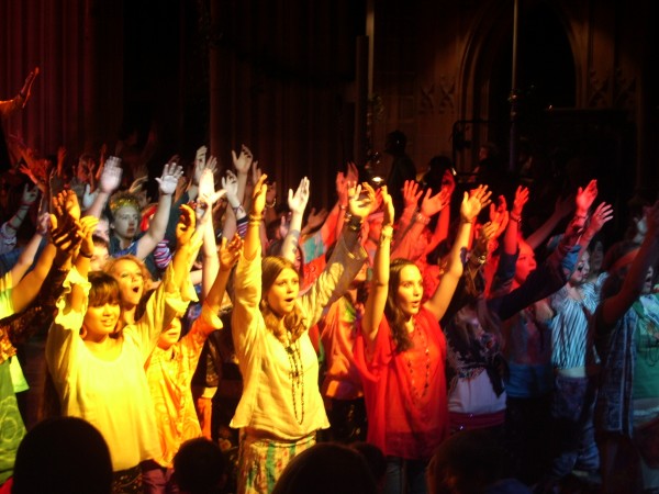 The chorus of Godspell raises their arms in the air.
